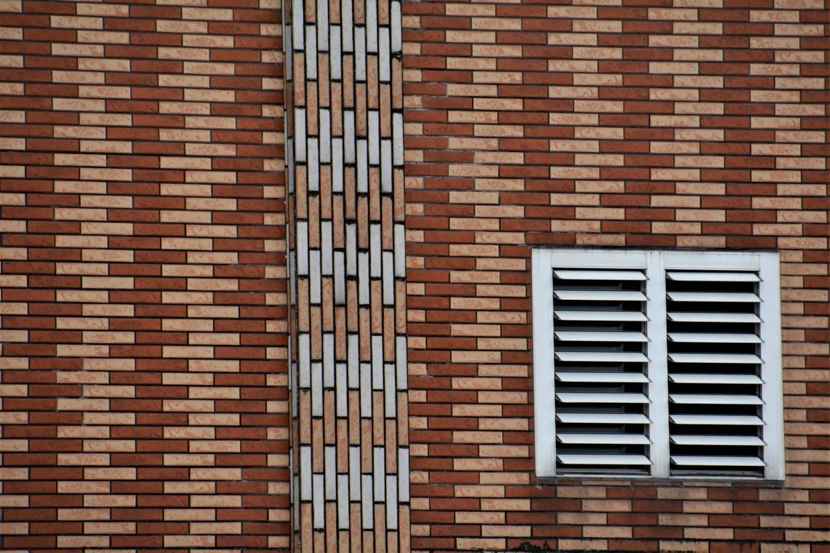 A white louvered vent set into a red brick wall, beside a vertical band of patterned bricks.