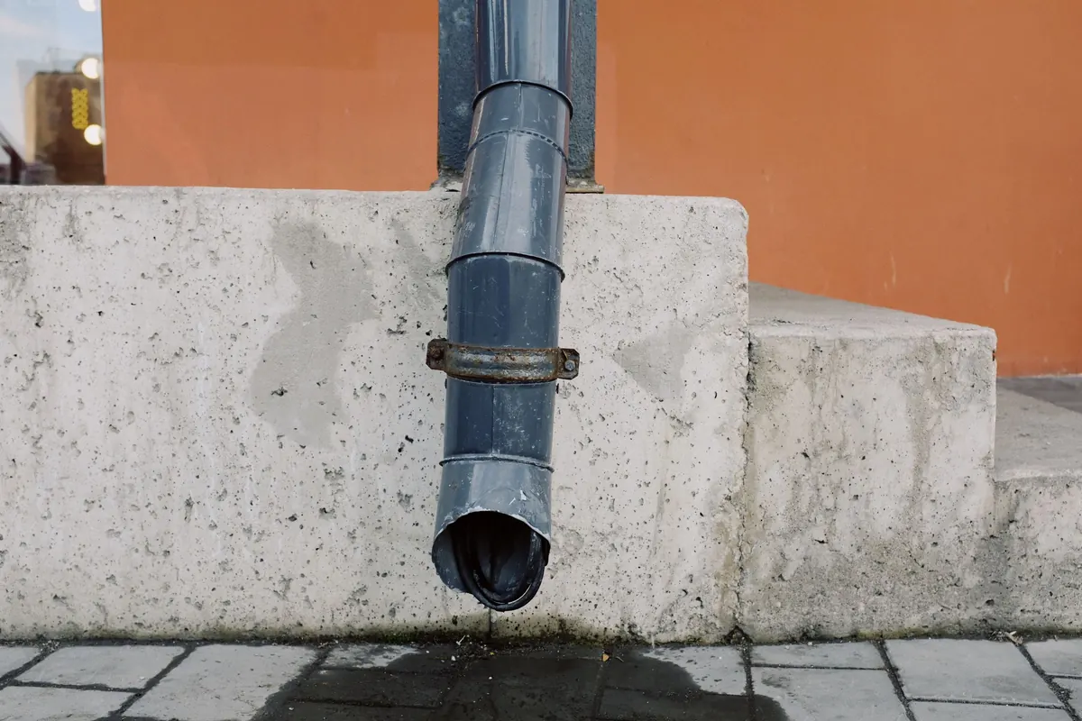 A gray downspout ending just above a concrete ledge at the base of a building, with a wet patch on the paving below.