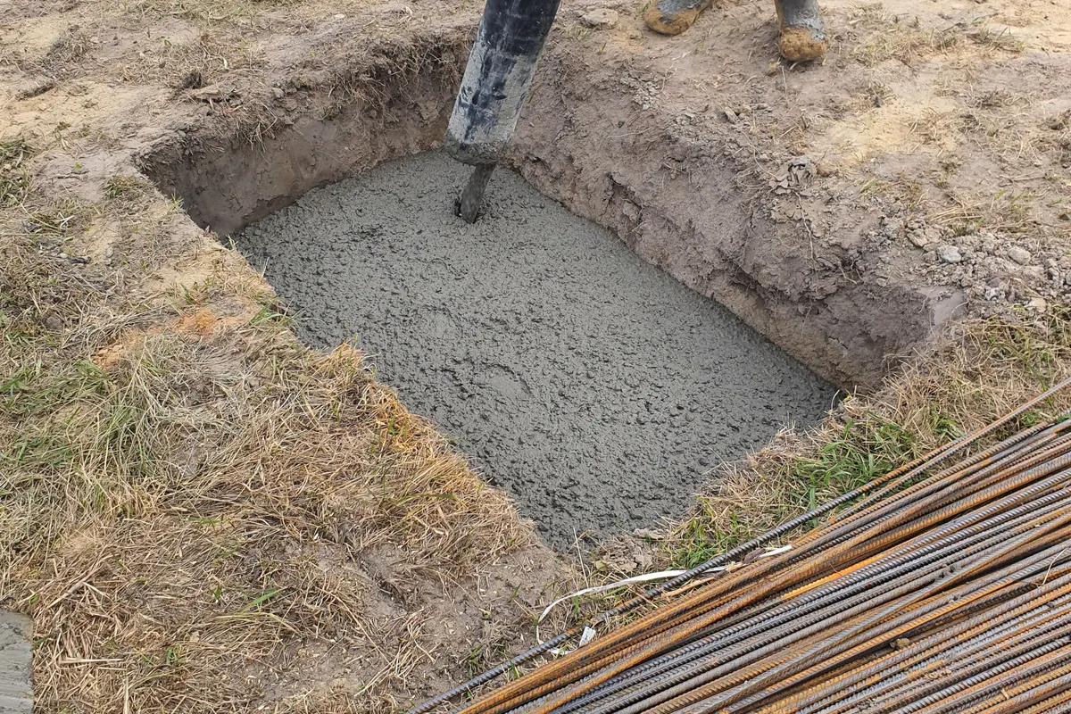 Concrete being poured from a hose into a square hole dug in the ground, with steel rebar lying beside it.